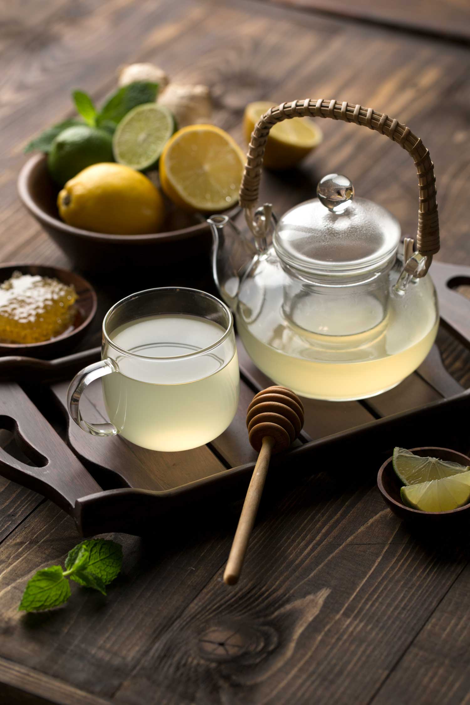 An image of a glass pot of tea and mug on wooden background half filled with a clear cloudy tea with lemons and honey in the background